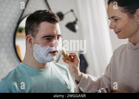 Mère appliquant de la mousse à raser sur le visage de son fils. Jeune homme trisomique apprenant à se raser. Banque D'Images