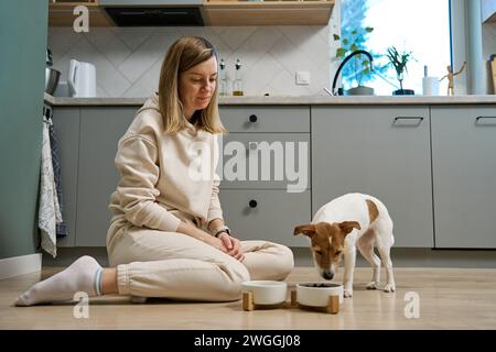 Femme mettant le bol de nourriture avec la nourriture pour son chien sur le sol dans la cuisine, propriétaire femme passant du temps avec l'animal de compagnie à la maison, alimentation animale et animal CA Banque D'Images