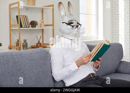 Jeune homme dans le masque drôle de lapin et des lunettes assis sur le canapé et lisant le roman Banque D'Images