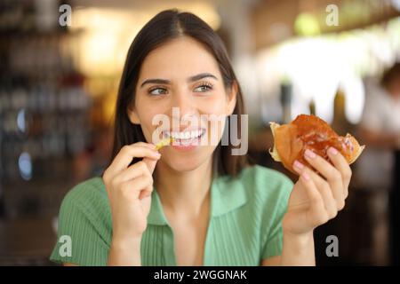 Femme heureuse mangeant hamburger et frites dans un café regardant de côté Banque D'Images