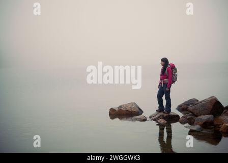 Une femme randonneuse se tient sur un rocher regardant au-dessus d'un étang Jordan enveloppé de brouillard dans le parc national Acadia dans le Maine. Banque D'Images