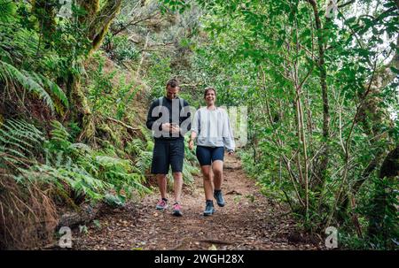 Un jeune couple sportif marche sur un sentier à travers la forêt en regardant leur téléphone Banque D'Images