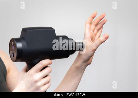Femme se donne massage à la main avec masseur-pistolet à percussion avec buse de balle. Gros plan. Fond de studio gris. Traitement pour soulager les douleurs articulaires. Banque D'Images