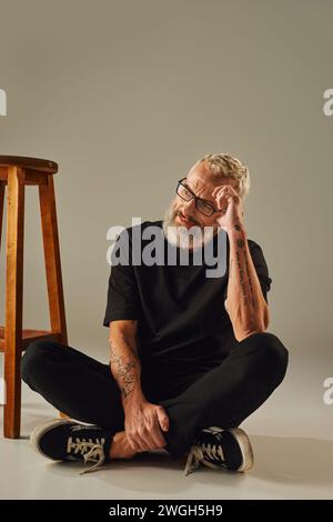 joyeux homme mûr beau en t-shirt noir avec tatouages posant à côté d'une grande chaise sur fond beige Banque D'Images