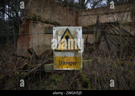 Un panneau d'avertissement de « danger » sale placé sur une clôture en treillis devant une maison détruite et abandonnée. Il y a des débris et des murs effondrés derrière elle. Banque D'Images