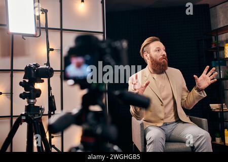 bel homme avec les cheveux roux et la barbe dans une tenue élégante parlant activement pendant podcast en studio Banque D'Images