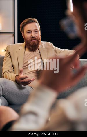 beau homme barbu avec les cheveux roux dans des vêtements élégants assis à côté de son intervieweur dans le studio Banque D'Images