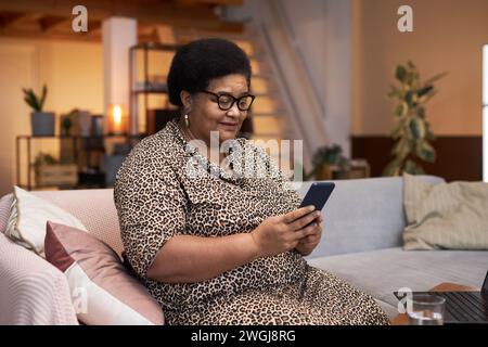 Portrait de femme senior noire moderne portant la robe imprimée guépard à l'aide d'un smartphone assis sur un canapé dans l'espace de copie intérieur de la maison Banque D'Images