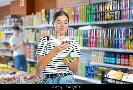 Fille scannant le code QR sur la canette de boisson gazeuse avec smartphone dans l'épicerie Banque D'Images