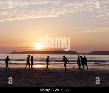 De jeunes hommes en silhouette jouent au football sur une plage de sable au coucher du soleil avec une île derrière à Essaouira, Maroc, le 5 février 2024 Banque D'Images