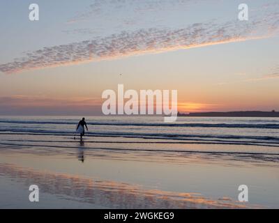 Surfeur en silhouette sort de la mer au coucher du soleil avec une île à l'horizon à Essaouira, Maroc, le 5 février 2024 Banque D'Images