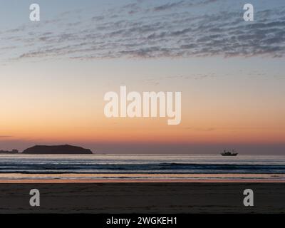 Le soleil se couche sur une île et un bateau de pêche reflétés dans l'eau, avec une plage de sable en premier plan à Essaouira, Maroc, le 5 février 2024 Banque D'Images