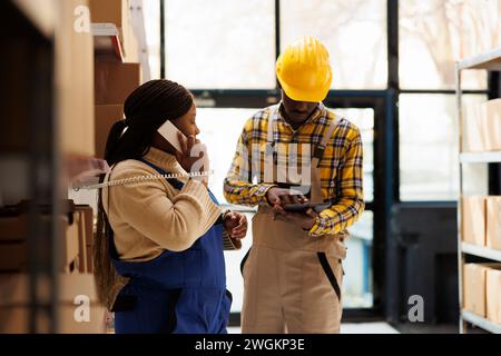 Employé de l'entrepôt vérifiant le colis dans la tablette et le gestionnaire répondant à l'appel du superviseur. Femme afro-américaine parlant sur le téléphone fixe et assistant à l'aide d'un logiciel d'inventaire dans l'entrepôt Banque D'Images