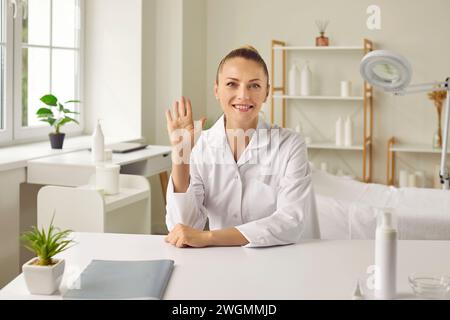 Femme heureuse esthéticienne assise à table, commençant la consultation en ligne de soins de la peau et saluant bonjour Banque D'Images