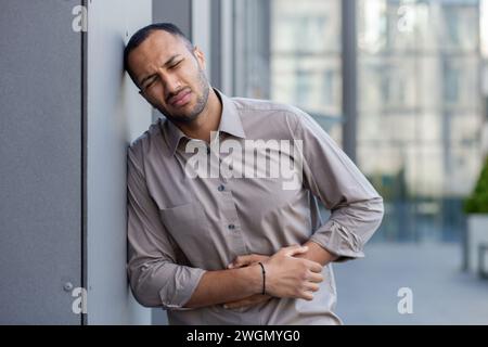 Mâle hispanique malade en vêtements de forme ayant mal au ventre et fermant les yeux dans la douleur tout en se tenant debout en ville. Homme d'affaires épuisé tenant les mains sur le ventre et souffrant de spasmes dus à une intoxication alimentaire. Banque D'Images