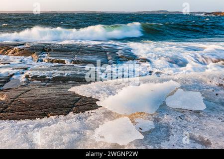 L'image capture la beauté dynamique des vagues qui se brisent sur un rivage rocheux recouvert de glace, illuminé par la douce lueur d'un soleil couchant. Banque D'Images