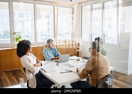 Une équipe de quatre professionnels est réunie autour d’une table blanche dans un bureau moderne, révélateur d’une réunion d’affaires coopérative et stratégique. Vue à travers la cloison de verre à une équipe de travail diversifiée Banque D'Images