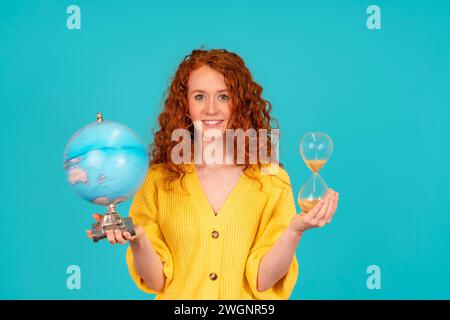 Femme joyeuse rousse en t-shirt jaune tenant le globe terrestre et Sandglass sur le concept d'éducation de fond bleu Banque D'Images