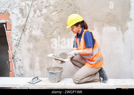 Ouvrière de construction féminine sur le mur de plâtrage de chantier de construction avec du ciment dans l'équipement de sécurité Banque D'Images