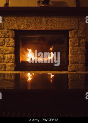 Vue rapprochée verticale en basse lumière d'une cheminée italienne dans une maison de campagne avec les flammes reflétées sur un verre de table basse. Sardaigne, Italie. Banque D'Images