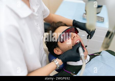Dentiste féminine utilisant le laser dentaire à diode moderne pour les soins parodontaux, vue de dessus. Patient de sexe masculin portant des lunettes de protection, empêchant les dommages à la vue. Banque D'Images