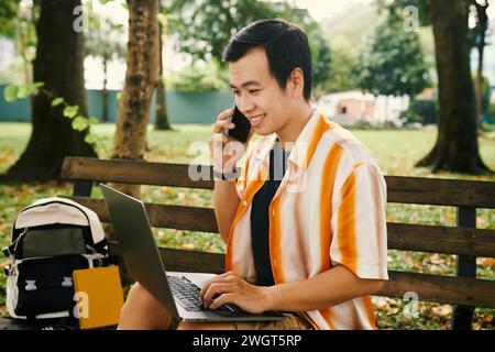 Jeune solopreneur masculin souriant avec smartphone par l'oreille tapant sur le clavier d'ordinateur portable et regardant l'écran tout en étant assis sur le banc dans le parc Banque D'Images