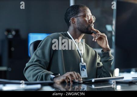 Vue de côté portrait d'un jeune homme noir portant des lunettes et pensant dans le bureau professionnel de l'entreprise regardant loin menton reposant sur l'espace de copie de la main Banque D'Images