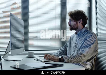 Portrait de vue latérale graphique de l'homme barbu à l'aide d'un ordinateur dans le bureau de l'entreprise INFORMATIQUE contre des fenêtres avec des stores sur l'espace de copie Banque D'Images