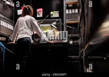 Femme afro-américaine détective privé regardant des photos de surveillance sur ordinateur portable et analysant des indices. Enquêteur debout près du bureau, étudiant les photographies et les preuves suspectes Banque D'Images