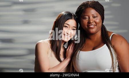 Joyeuses dames interraciales riant de la caméra et s'embrassant, appréciant l'amitié et exprimant la positivité corporelle. Jeunes femmes se sentant confiantes et belles, skinny et courbes types de corps. Banque D'Images