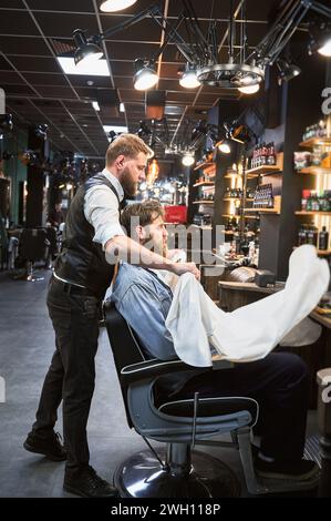 Coiffeur de fantaisie mettant une cape sur le client. Barbier professionnel préparant tout pour rendez-vous. Homme confiant visitant un salon de coiffure. Banque D'Images
