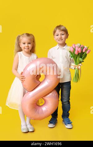 Petits enfants mignons avec ballon à air rose en forme de figure 8 et bouquet de belles tulipes sur fond jaune. Journée internationale de la femme Banque D'Images