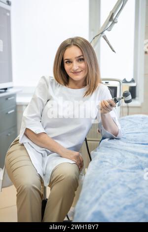 Jolie femme dermatologue avec dermatoscope en uniforme et gants dans le cabinet du médecin, salle d'examen en clinique. Jeune spécialiste Banque D'Images