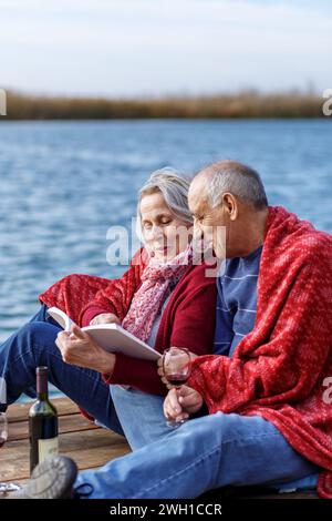 Heureux couple senior profitant du temps ensemble lisant un livre au bord du lac envelopper dans une couverture rouge. Banque D'Images