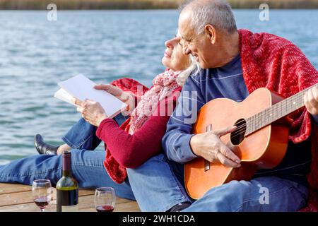 Heureux couple senior profitant du temps ensemble jouer de la guitare et boire du vin au bord du lac envelopper dans une couverture rouge. Banque D'Images