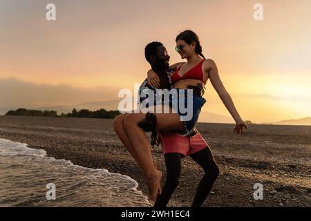 Joyeux couple interracial embrassant au bord de la mer, partageant un moment de lumière sur un magnifique coucher de soleil en toile de fond. Banque D'Images