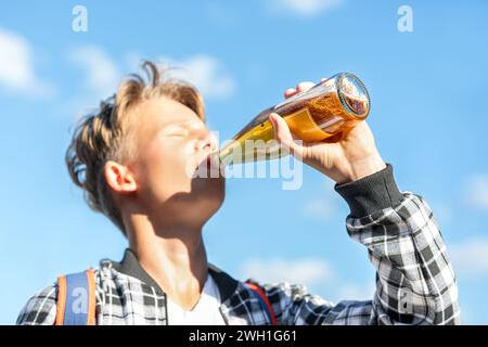 Joyeux garçon buvant de la limonade dans une bouteille en verre contre un beau ciel en toile de fond. Il porte une chemise à carreaux et un sac à dos. Couleurs éclatantes Banque D'Images