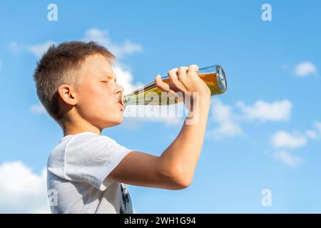 Joyeux garçon en T-shirt blanc buvant de la limonade d'une bouteille en verre sur un magnifique fond de ciel. Prise de vue en angle latéral. Couleurs éclatantes Banque D'Images