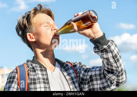 Joyeux garçon buvant de la limonade dans une bouteille en verre contre un beau ciel en toile de fond. Il porte une chemise à carreaux et un sac à dos. Couleurs éclatantes Banque D'Images