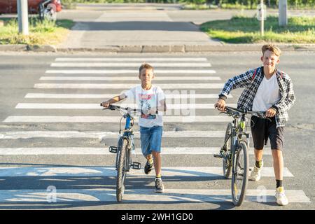 Deux garçons en tenue de tous les jours marchant et roulant des vélos à travers un passage pour piétons, adhérant aux règles de passage à niveau en tant que cyclistes Banque D'Images