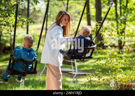 La mère avec deux enfants passe un moment amusant à l'aire de jeux. La mère balançait ses enfants sur les balançoires. Banque D'Images