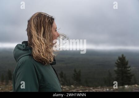 Vue de face latérale d'une femme aux cheveux blonds soufflant dans le vent dans la nature nordique, à l'esprit regardant loin au-dessus de la forêt et les nuages orageux en arrière-plan Banque D'Images