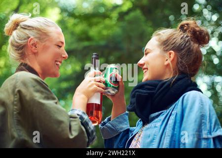 Amis, acclamations et boissons au festival de musique en forêt, excité et bière pour se lier ensemble par les arbres. Femmes, boire et célébrer en été Banque D'Images