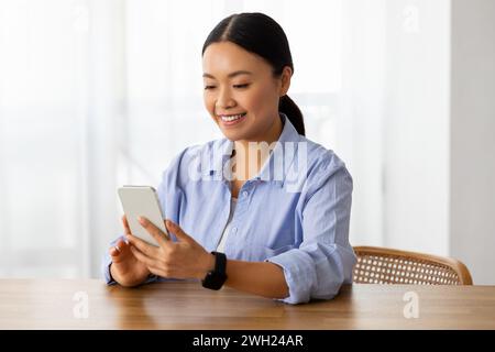 Femme asiatique souriante assise au bureau à la maison, en utilisant le téléphone Banque D'Images