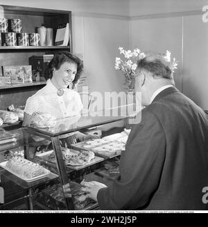 La pâtisserie dans les années 1950 Une pâtisserie à Eskilstuna où un client masculin se tient debout et achète au comptoir bien approvisionné avec des pâtisseries, des brioches et des gâteaux. Une jeune femme travaille dans la pâtisserie et a l'air heureuse en racontant au client masculin la sélection des pâtisseries. 3 juin 1954. Conard ref 2687 Banque D'Images