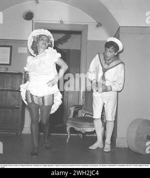 Un jeune couple aux vêtements bizarres dans les années 1940 Elle est habillée de blanc avec des boucles dans ses cheveux et a soulevé sa jupe pour que ses jambes soient pleinement visibles. Les bas hauts sont attachés aux porte-jarretières. L'homme en costume de marin se tient debout et regarde. Deux acteurs dans leurs costumes de scène en coulisses. 1944. Kristoffersson réf. K134-5 Banque D'Images