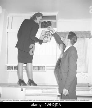 Un couple dans la cuisine dans les années 1940 Probablement dans leur nouvelle maison, ils ont mis des paquets de café et de détergent dans les placards de la cuisine. Elle se tient debout sur le comptoir de la cuisine avec ses chaussures pour atteindre le placard de la cuisine. L’homme lui tend un sac en maille plein d’emballages avec des marques classiques telles que RADION, Omo, Sunlight Two et Vim. 1947. Kristoffersson ref AB7-2 Banque D'Images