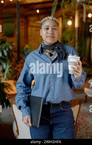 Portrait vertical de femme noire portant une tenue en denim dans un bureau de coworking vintage et tenant un ordinateur portable Banque D'Images