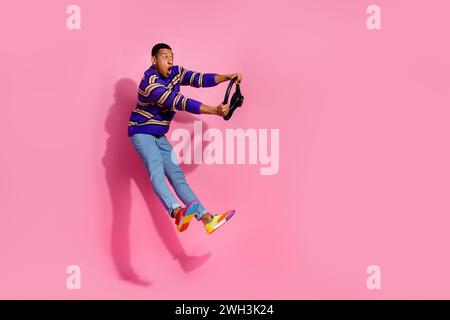 Photo du corps entier de jeune homme attrayant saut dangereux conduire porter des vêtements à rayures violettes à la mode isolés sur fond de couleur rose Banque D'Images