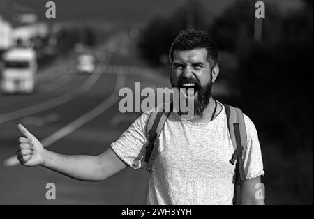 Arrêt automatique du déplacement. Taille basse essayez d'arrêter la voiture et le pouce vers le haut geste. Homme au visage joyeux et à la barbe voyageant en randonnée avec la route en arrière-plan Banque D'Images